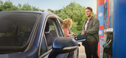 Person refueling a blue car at a gas station while smiling and talking with a child leaning out of the back window.