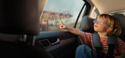Child in a car seat drawing smiley faces on a fogged-up window.