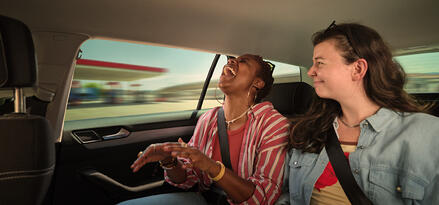 Two friends laughing and chatting in the back seat of a car during a road trip.