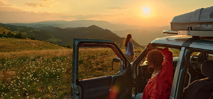 Two women pause during a road trip, one sitting in an SUV and the other standing in a wildflower-covered field, overlooking mountains at sunset.